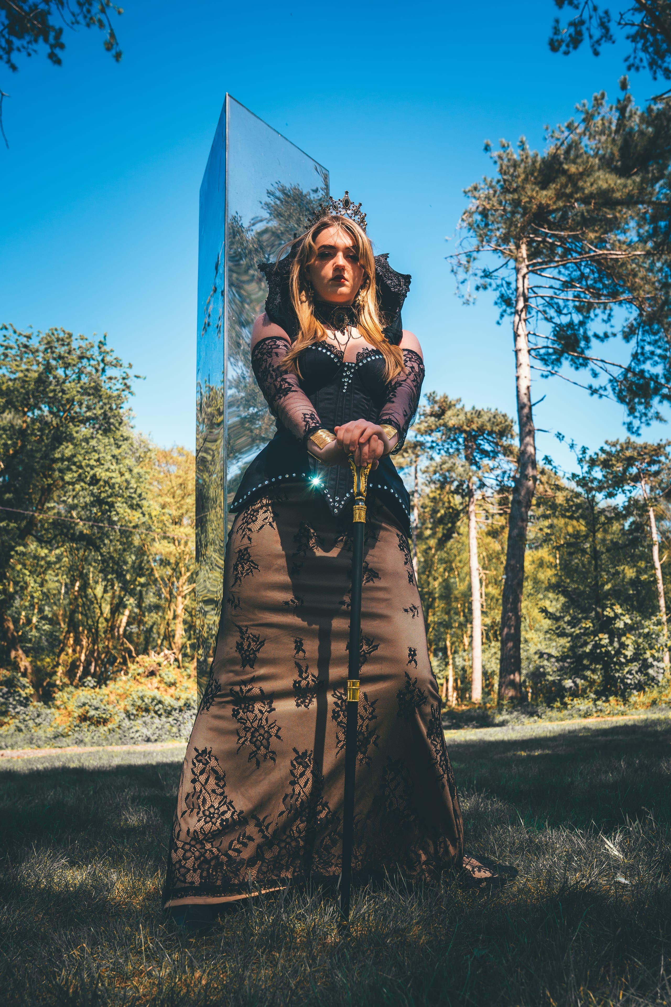 The Empress, guardian of the Castle Realm: a dark crown, a black corseted gown and gold gauntlets on a staff, photographed in the pines.