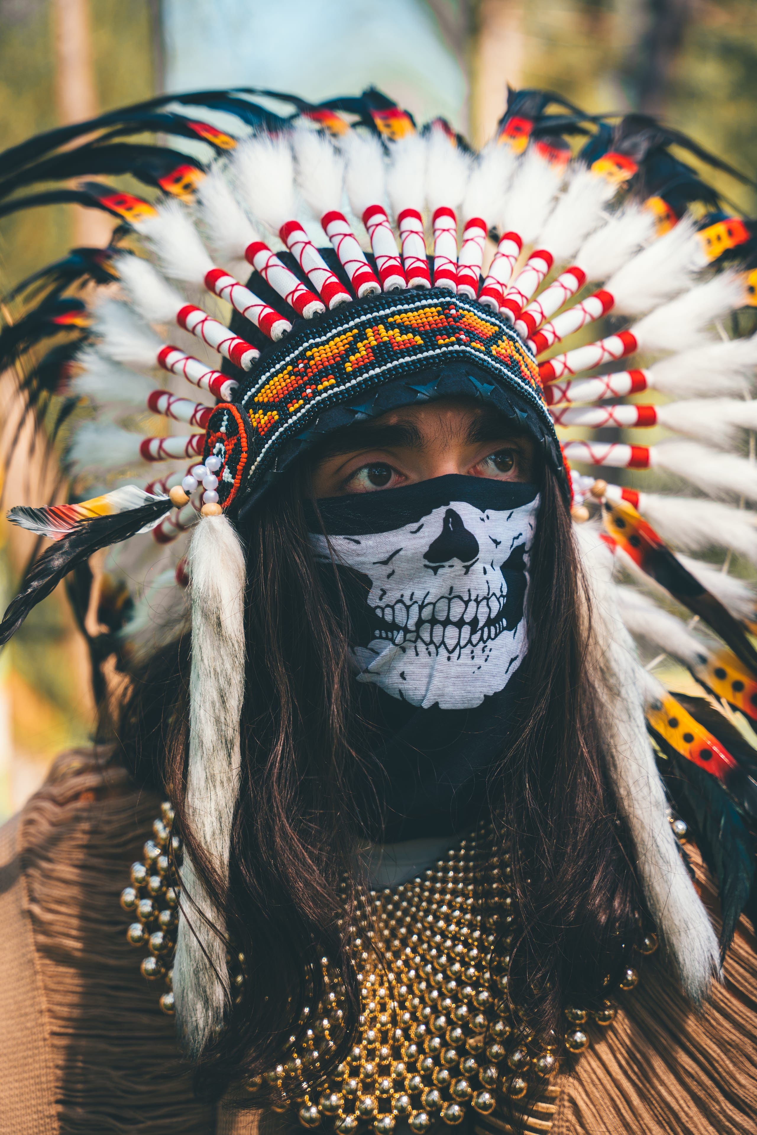 The Oracle, guardian of the Pyramid Realm: a feathered headdress and a skull-painted face covering, photographed in the pines.