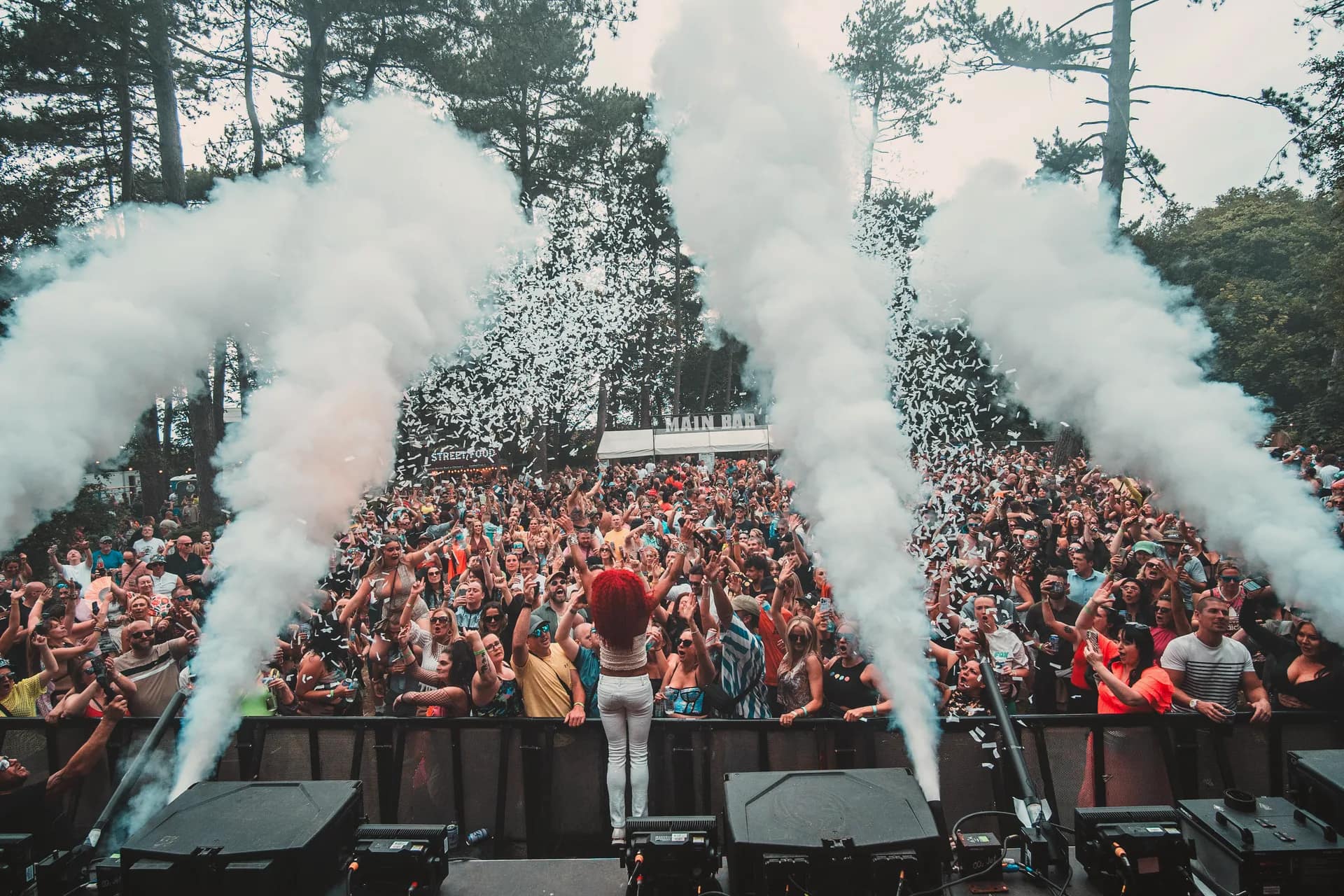 Confetti and smoke firing over the crowd in daylight, the main bar and street food stalls behind them