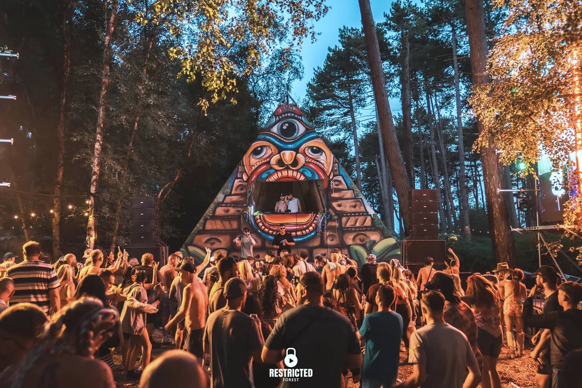 The totem pyramid stage at golden hour, festoon lighting through the pines and the crowd dancing in front of it.