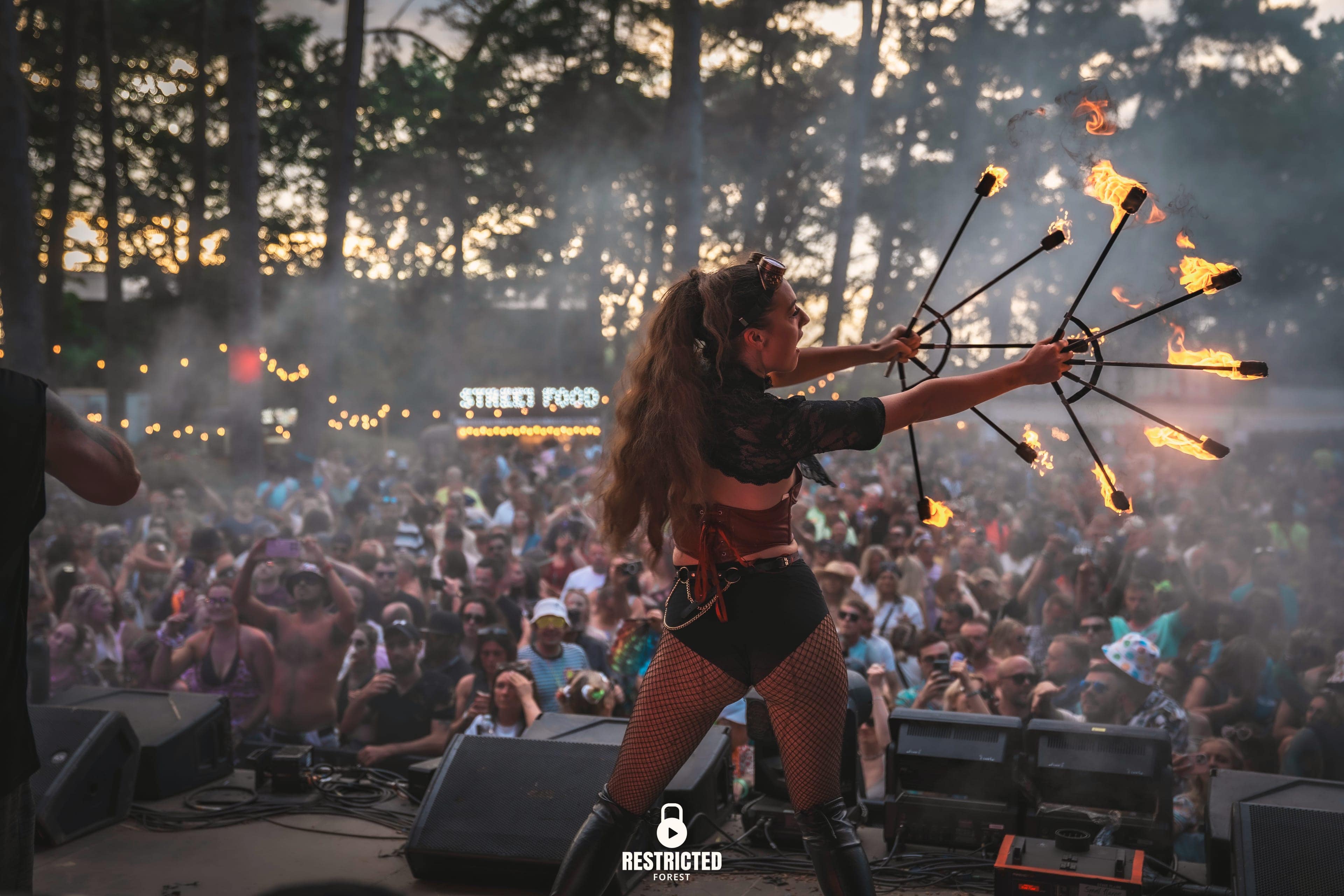 A fire performer seen from behind on the stage at golden hour, fire fans spread wide, smoke drifting through the pines and the packed crowd facing her, the Street Food sign lit at the back of the clearing