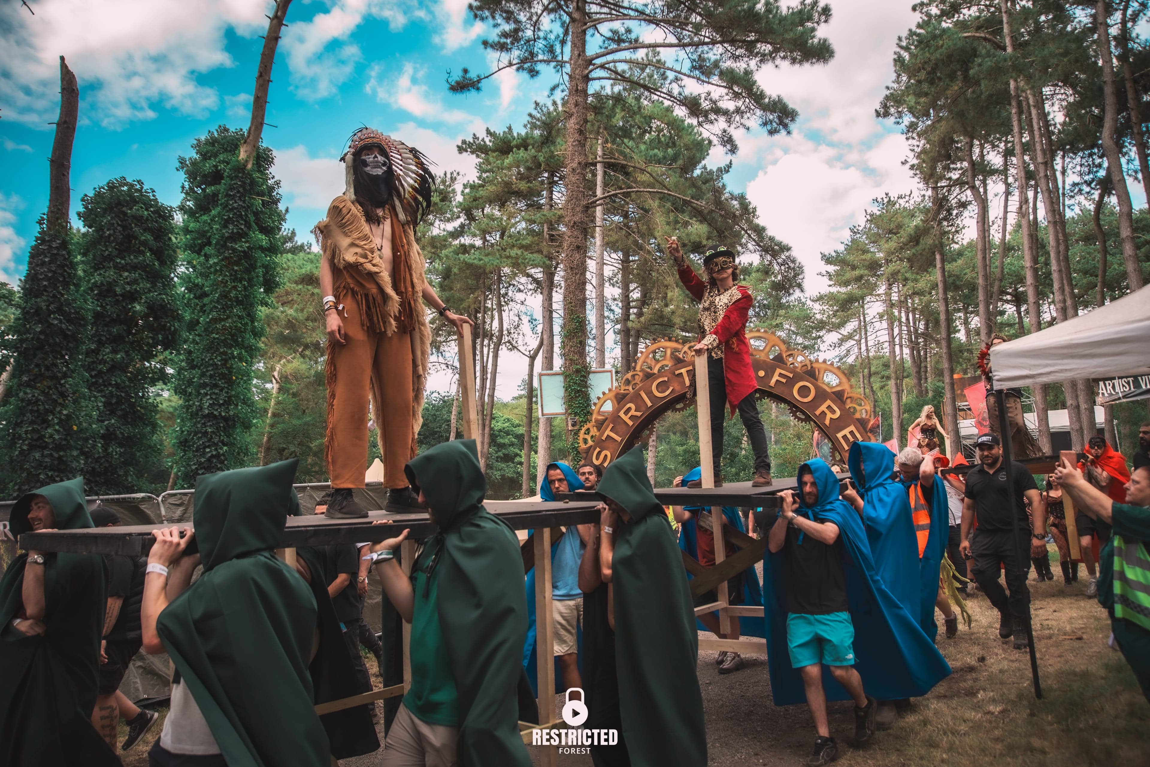 The procession: two figures carried above the crowd on platforms by green and blue cloaked bearers, the cog arch behind. Source frame RestrictedForest2026-1494.