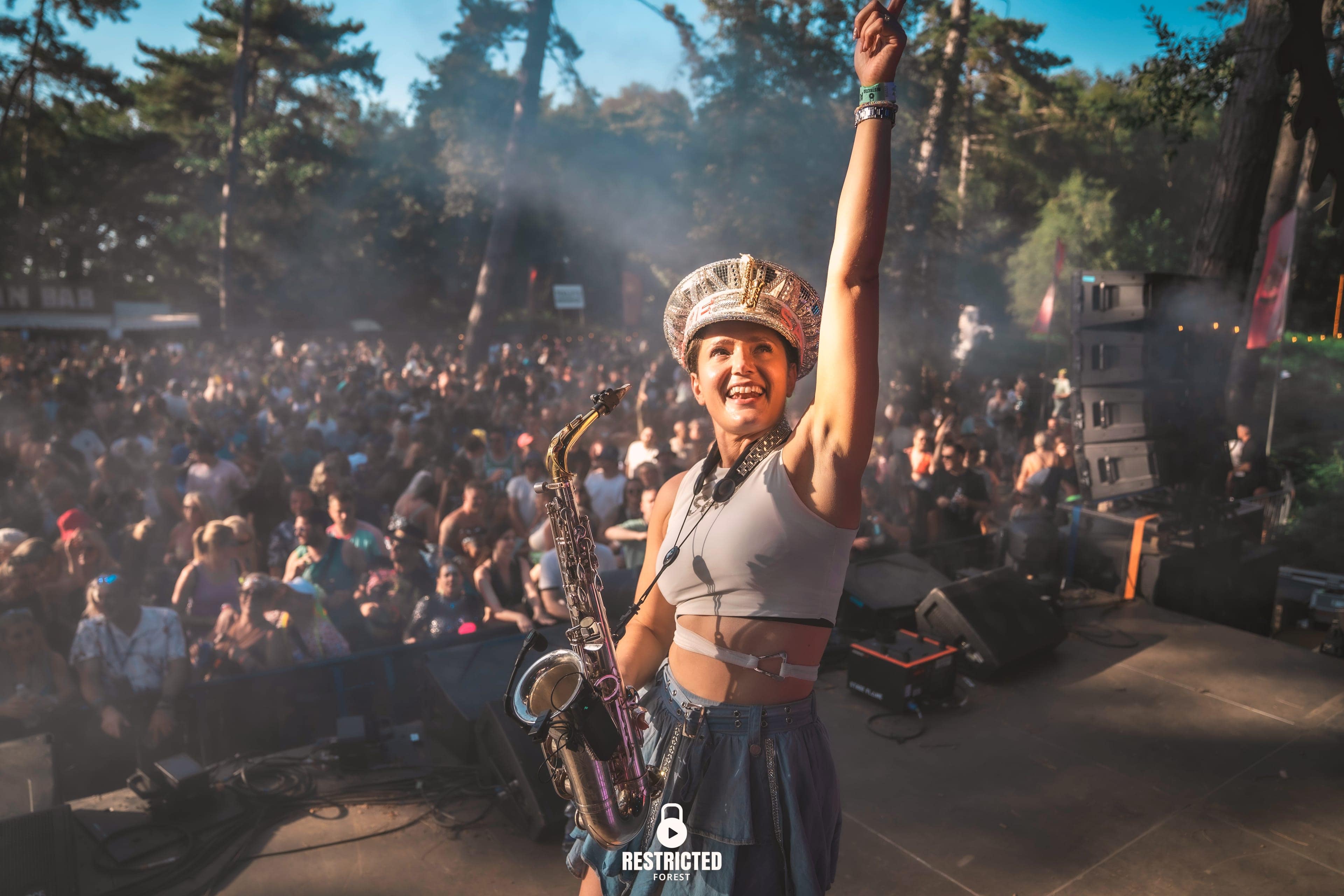 A saxophonist in a jewelled cap on stage in daylight, one arm raised to the crowd filling the clearing behind her